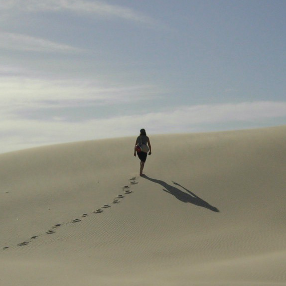 Frau geht über eine Düne, links sind Fußspuren, rechts ihr Schatten zu sehen.